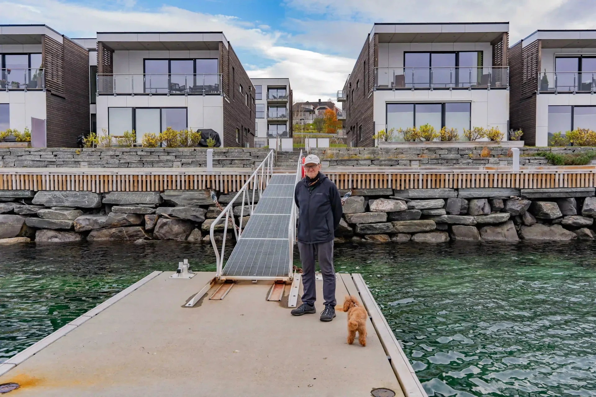 Kjell og hunden står på flytebrygga foran Havspeil. Bak dem ses landgangen, steinmuren mot sjøen og leilighetene med store vinduer og glassrekkverk.