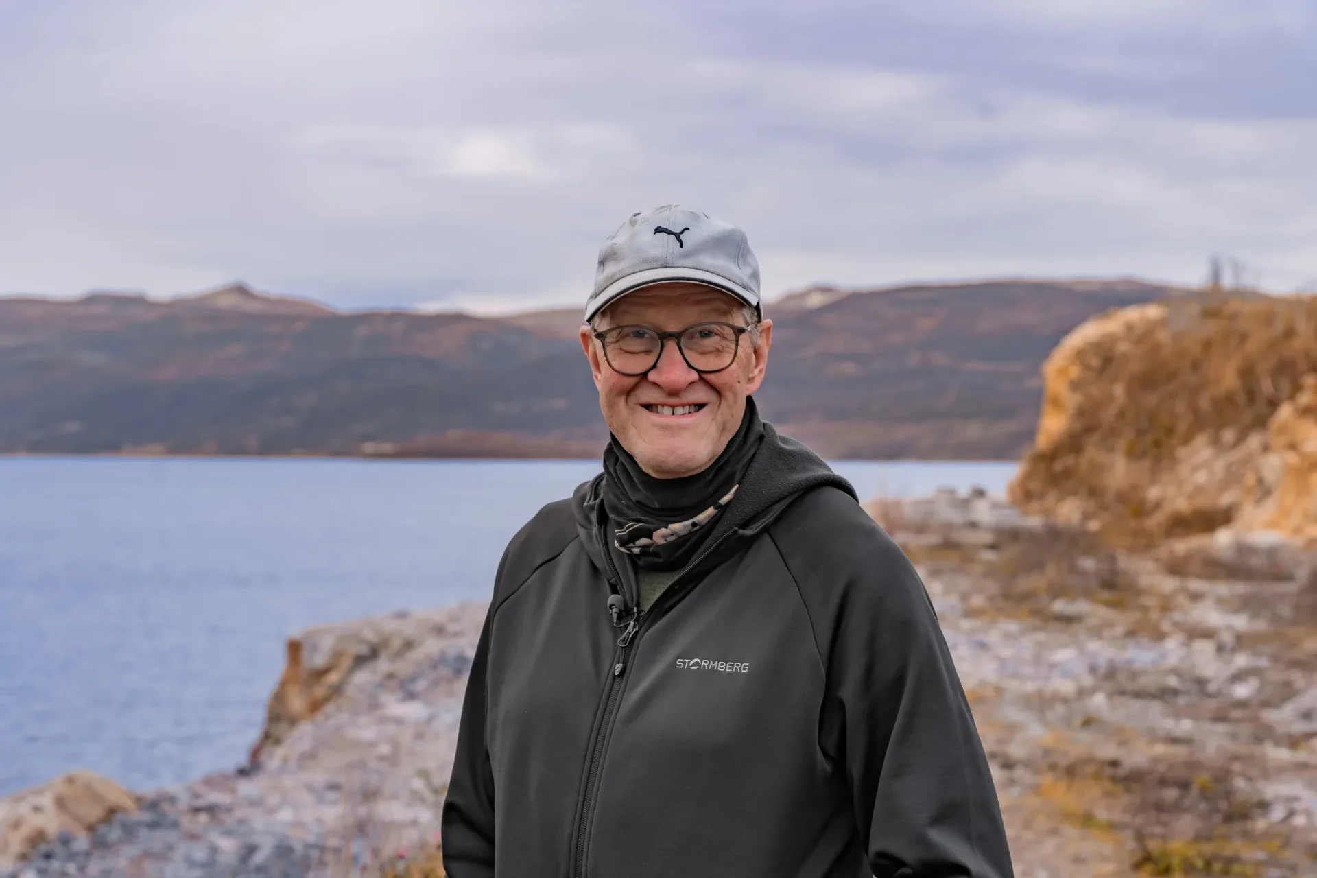 Portrett av Kjell med caps og briller, smilende foran Altafjorden og fjellene på andre siden.