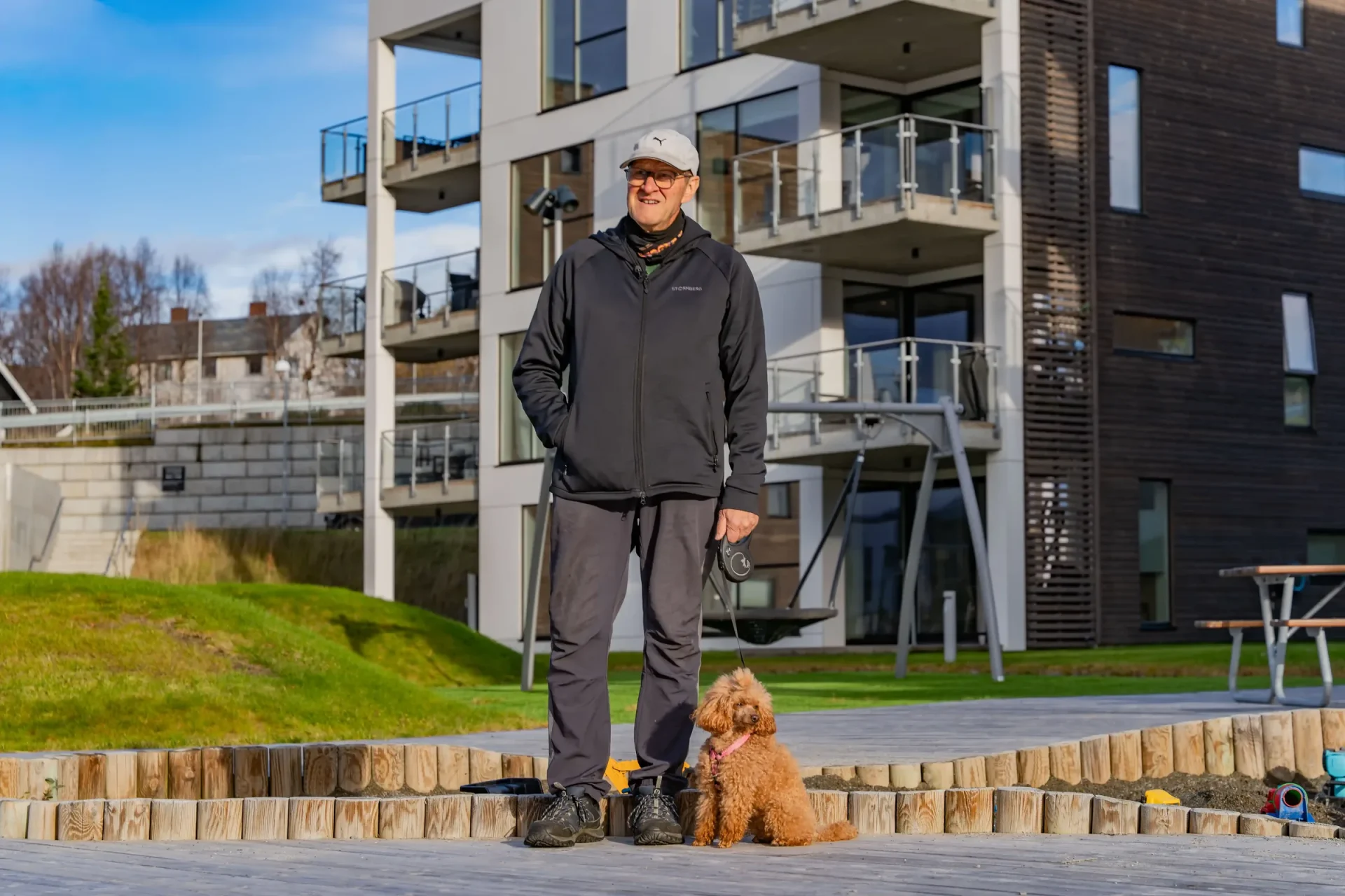Kjell står smilende med en liten brun puddel i bånd på uteområdet mellom leilighetsbyggene. Bak dem ses sandkassen, en rund huske og et bord med benker.