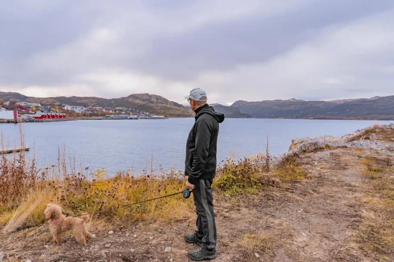 Kjell står med ryggen til og ser utover Altafjorden mot Bossekop, med en liten brun puddel i bånd ved siden av seg.